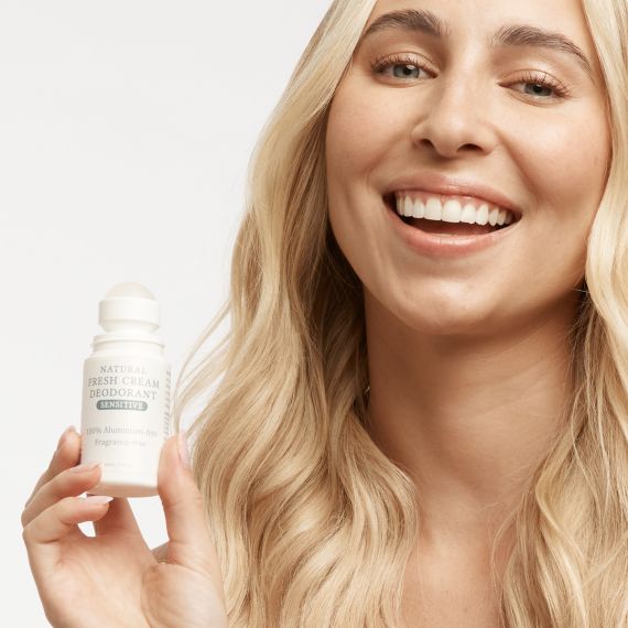 Smiling female model with long blonder hair holding the MooGoo Fresh Cream Deodorant in Sensitive with lid off and roller ball exposed. 