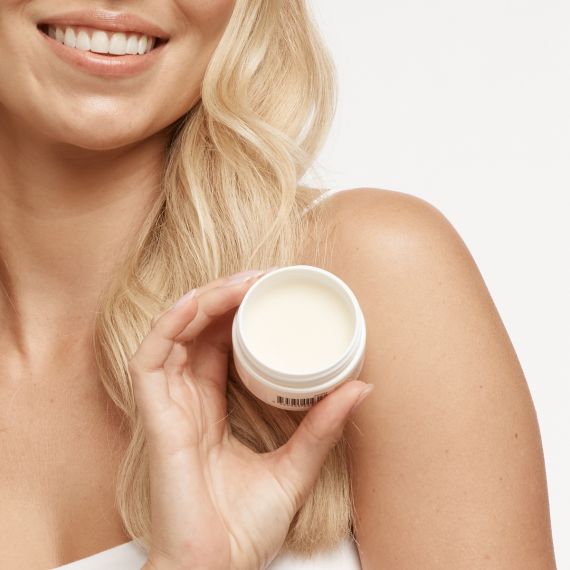 Smiling blonde haired model holding the MooGoo Natural Shea Sorbet Vanilla Butter Balm in front of her left shoulder with the lid off. 