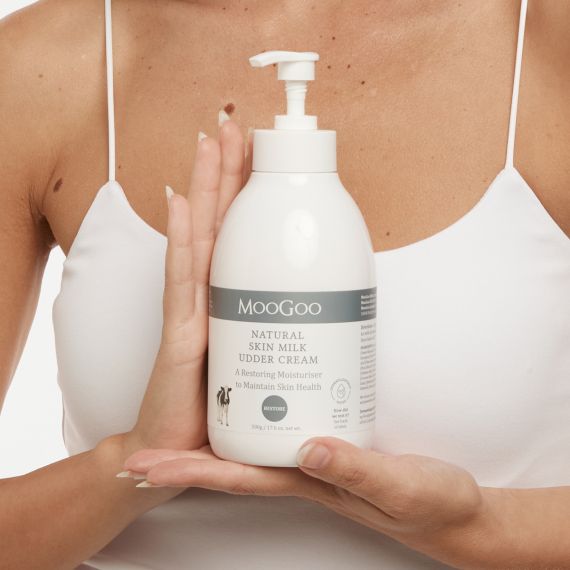 Female model wearing white singlet top holding the MooGoo Natural Skin Milk Udder Cream bottle in front of her chest with both hands. 