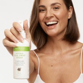 Smiling female model with olive skin and short brown hair holding the MooGoo Natural Tail Swat Spray up to the camera with her right hand. 