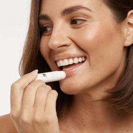 Smiling female model with short brown hair applying the MooGoo Natural Coconut Lip Balm to her lips. 