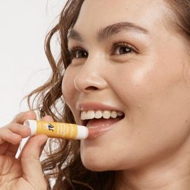 Smiling female of mixed islander and western heritage with curly brown hair and brown eyes applying the MooGoo Natural Tingling Honey Lip Balm from a side angle. 