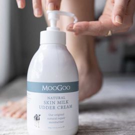 MooGoo Natural Skin Milk Udder Cream 500g on a bedroom floor with legs in the background and hands reaching down to pump the bottle. 